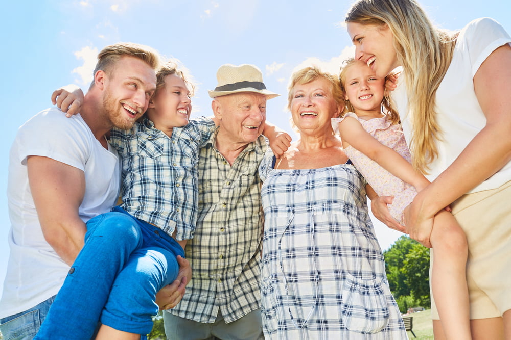A happy multi-generational family stands together outdoors, smiling and embracing under a bright blue sky - Primary Care Sheridan WY